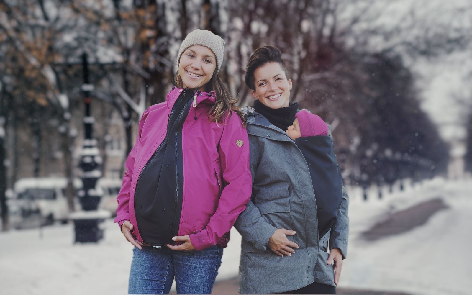 A pregnant woman and a babywearing woman standing side by side smiling with jacket extenders on their jackets in a snowy park 