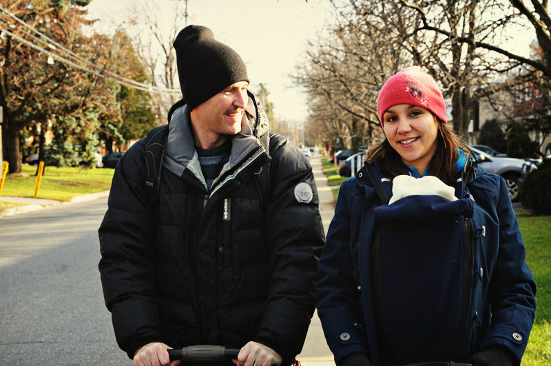 Husband and wife out walking down a suburb sidewalk. Husband is pushing a stroller and smiling at his wife who is baby wearing with a jacket extender.